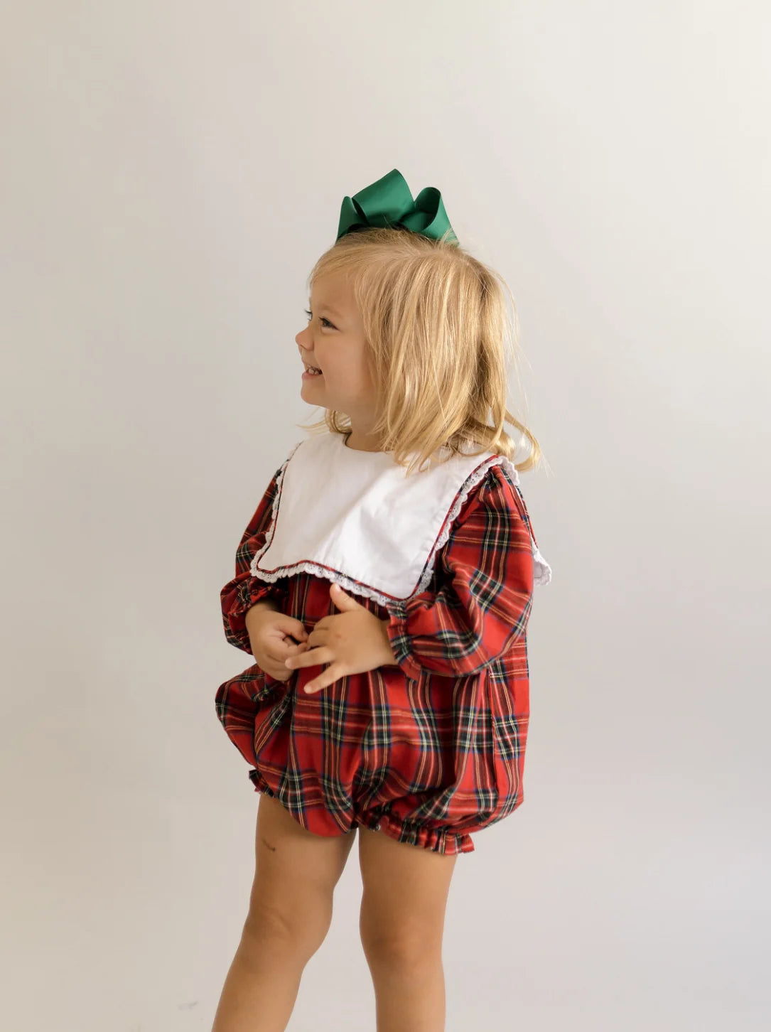 Toddler in red and green plaid bubble romper smiling, side view