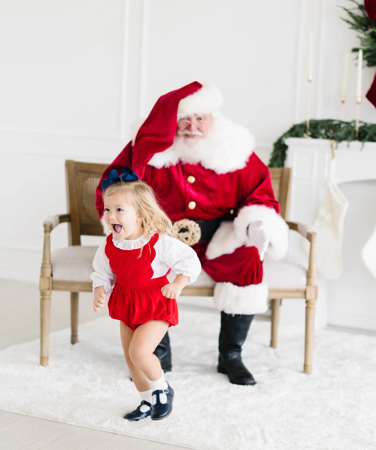 Toddler in red corduroy bubble romper with navy bow, running near Santa