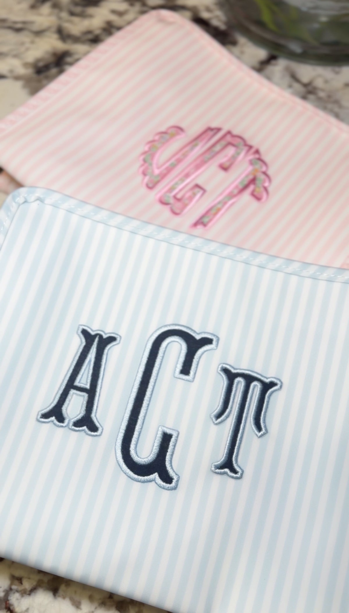 Blue and white striped TRVL Roadie pouch embroidered with 'ACT' monogram in navy serif letters, shown next to a pink monogrammed pouch