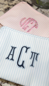 Blue and white striped TRVL Roadie pouch embroidered with 'ACT' monogram in navy serif letters, shown next to a pink monogrammed pouch