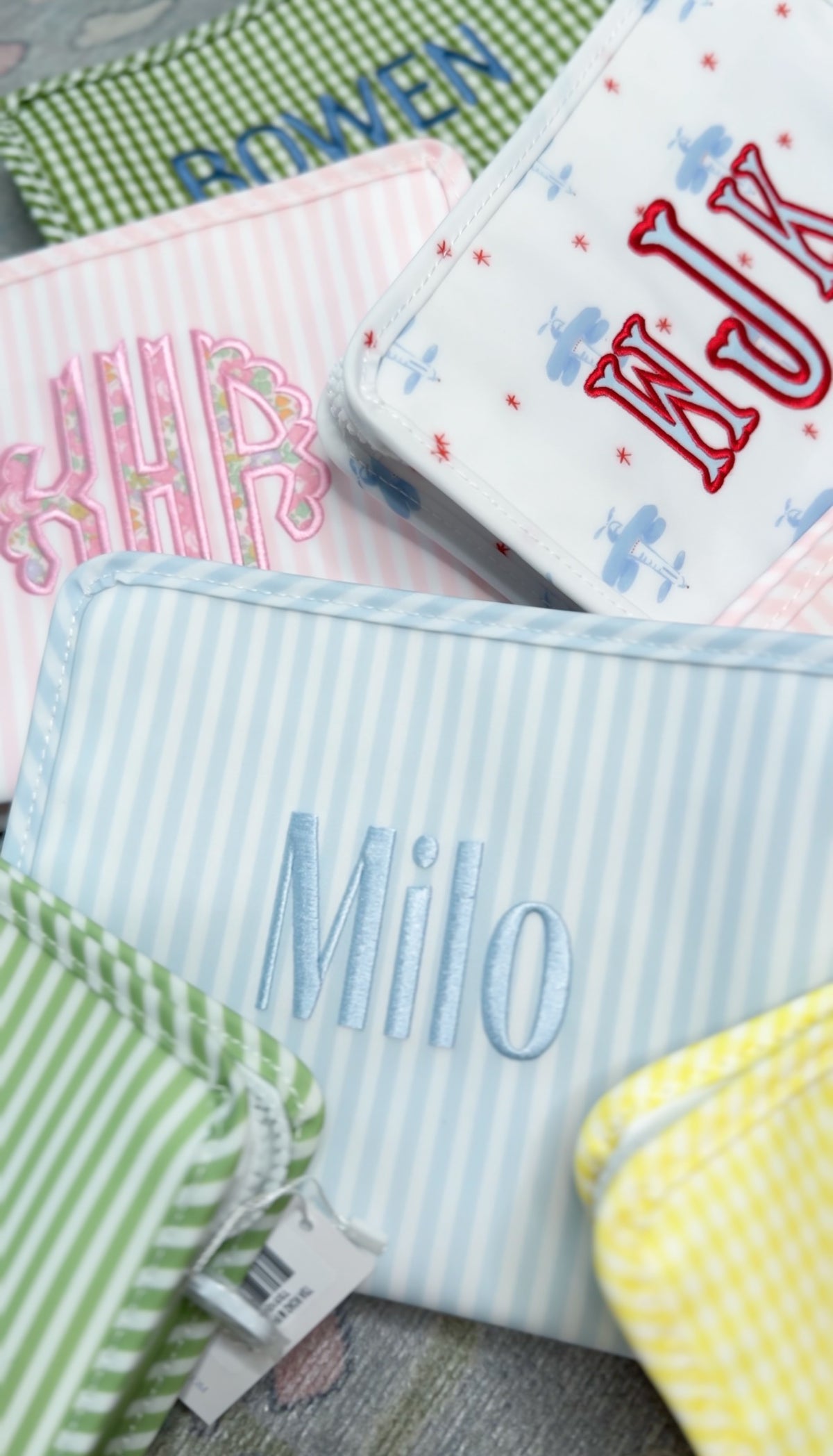 Blue and white striped TRVL Roadie pouch embroidered with 'Milo', shown alongside other monogrammed pouches in various colors