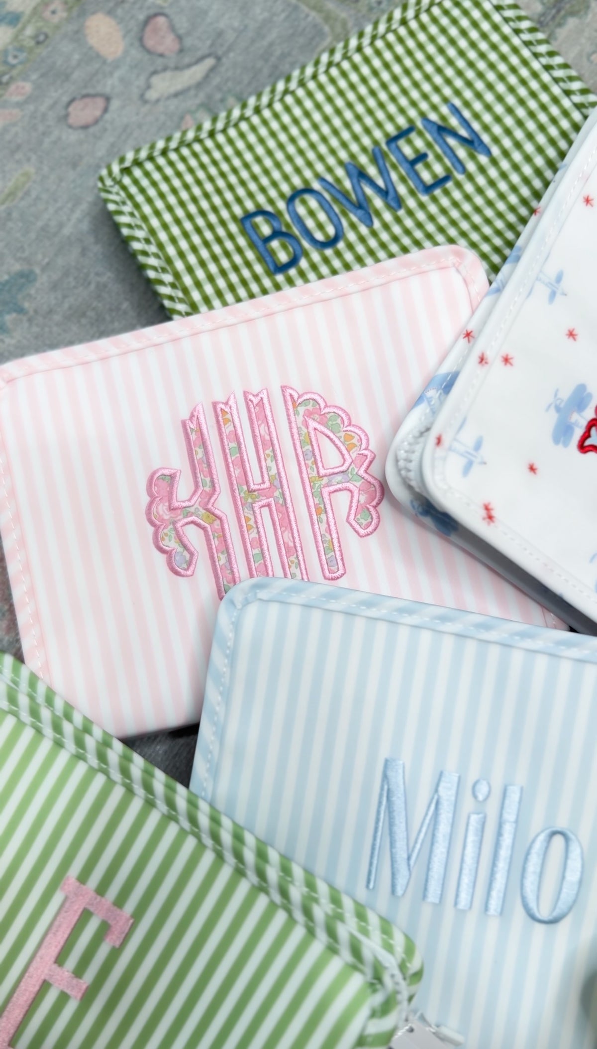 Pink Pimlico stripe TRVL Roadie pouch embroidered with 'KHP' monogram in floral-style letters, shown alongside other monogrammed pouches