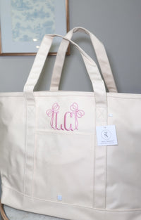 A beige natural coated canvas maxi tote with two handles, a front pocket, and a brand tag attached to it.