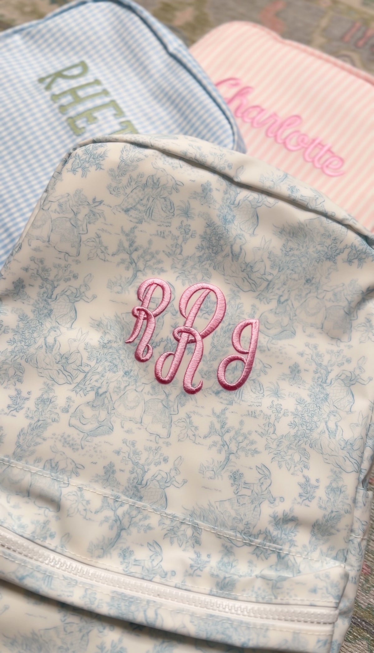 Cream bunny toile TRVL Backpacker embroidered with the pink 'RRJ' monogram, next to blue gingham and pink striped bags
