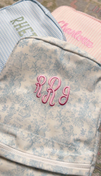 Cream bunny toile TRVL Backpacker embroidered with the pink 'RRJ' monogram, next to blue gingham and pink striped bags