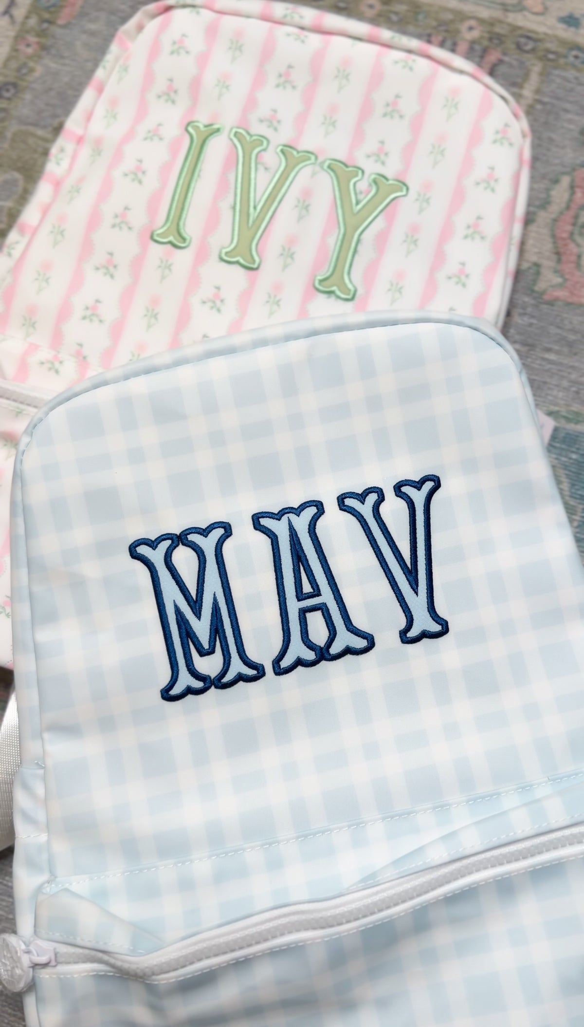 Light blue plaid TRVL Backpacker embroidered with the navy 'MAV' monogram, next to a pink striped bag embroidered 'Ivy'