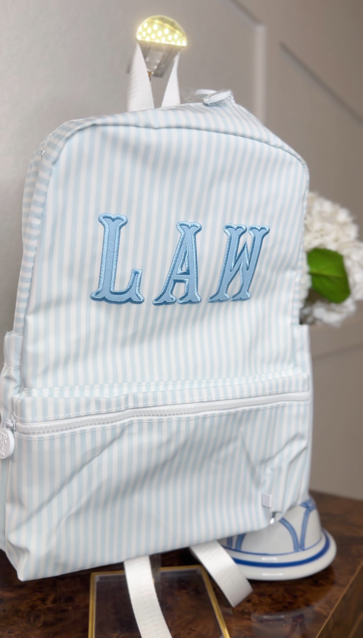 Light blue and white striped TRVL Backpacker on a stand, embroidered with the blue 'LAW' monogram
