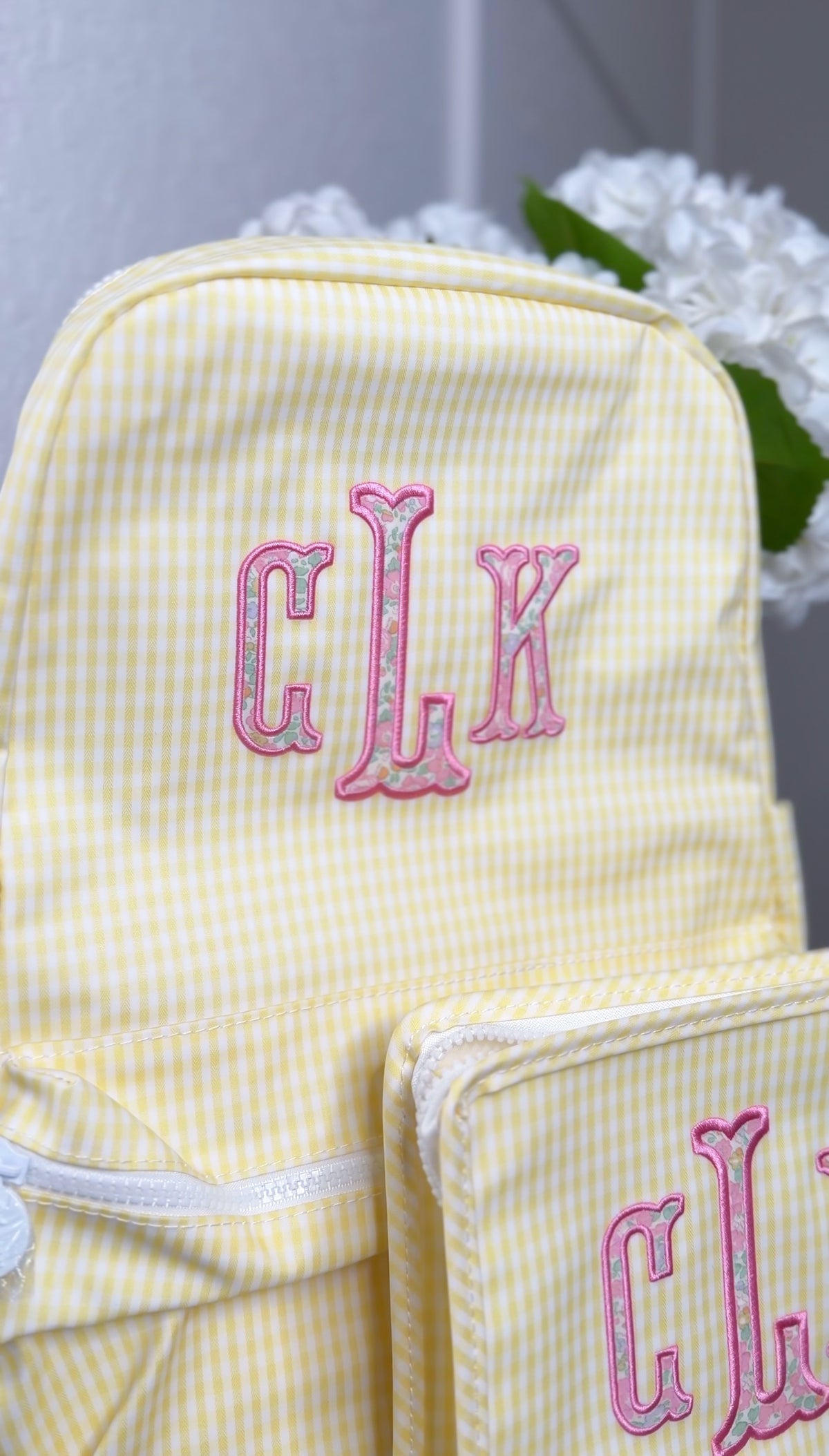 Yellow gingham TRVL Backpacker with pink embroidered 'CLK' monogram, matching bag visible beside it