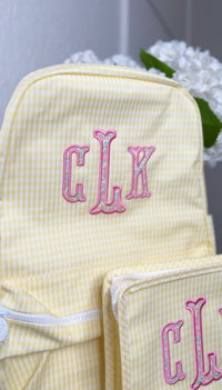 Yellow gingham TRVL Backpacker with pink embroidered 'CLK' monogram, matching bag visible beside it