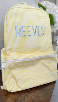 Yellow gingham TRVL Backpacker on a table with blue embroidered name 'Reeves'