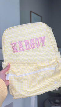 Woman holding a yellow gingham TRVL Backpacker with pink embroidered name 'Margot'