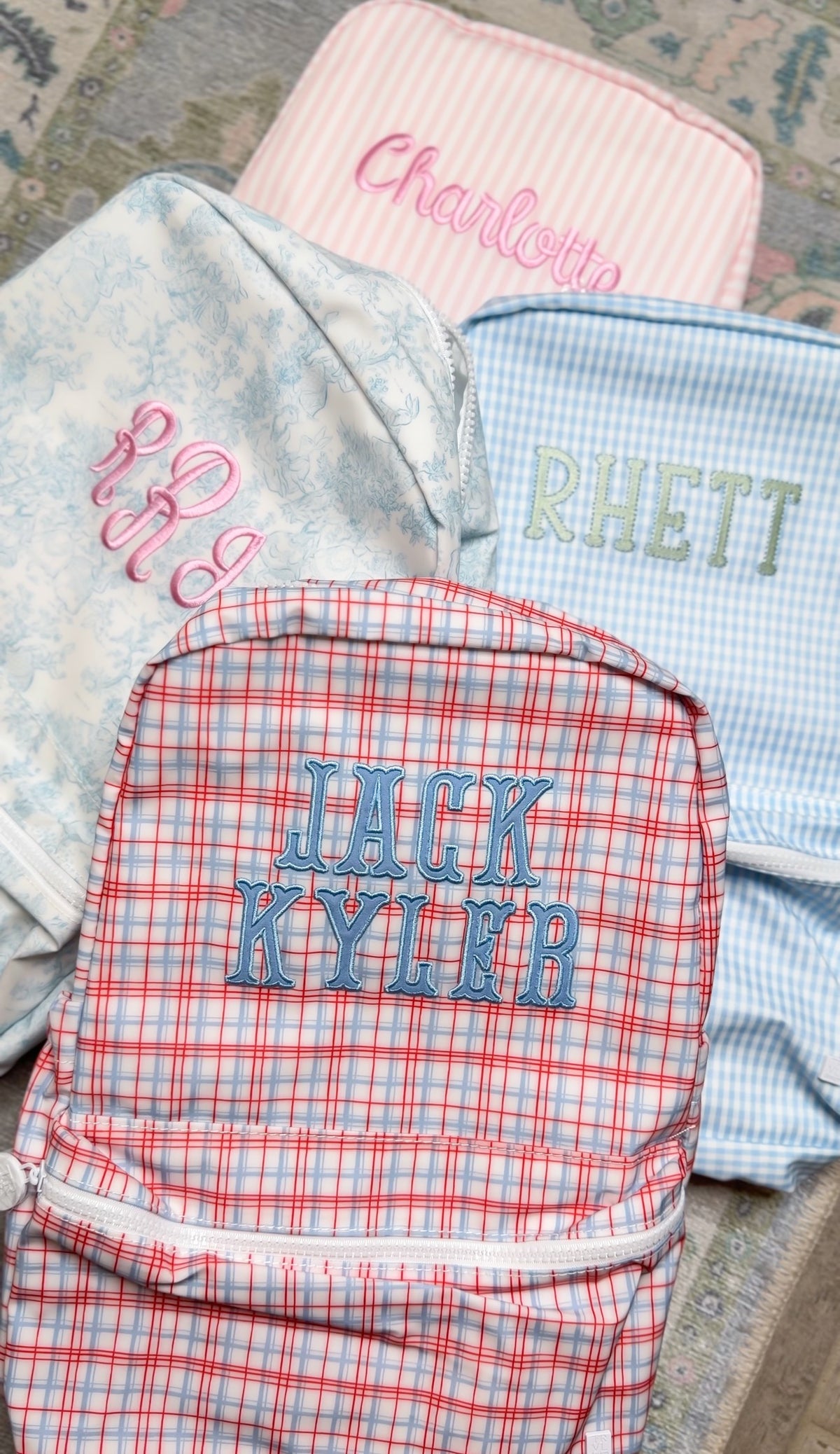 Red plaid TRVL Backpacker embroidered 'Jack Kyler', shown with pink striped, floral, and blue gingham bags in the background
