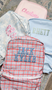 Red plaid TRVL Backpacker embroidered 'Jack Kyler', shown with pink striped, floral, and blue gingham bags in the background