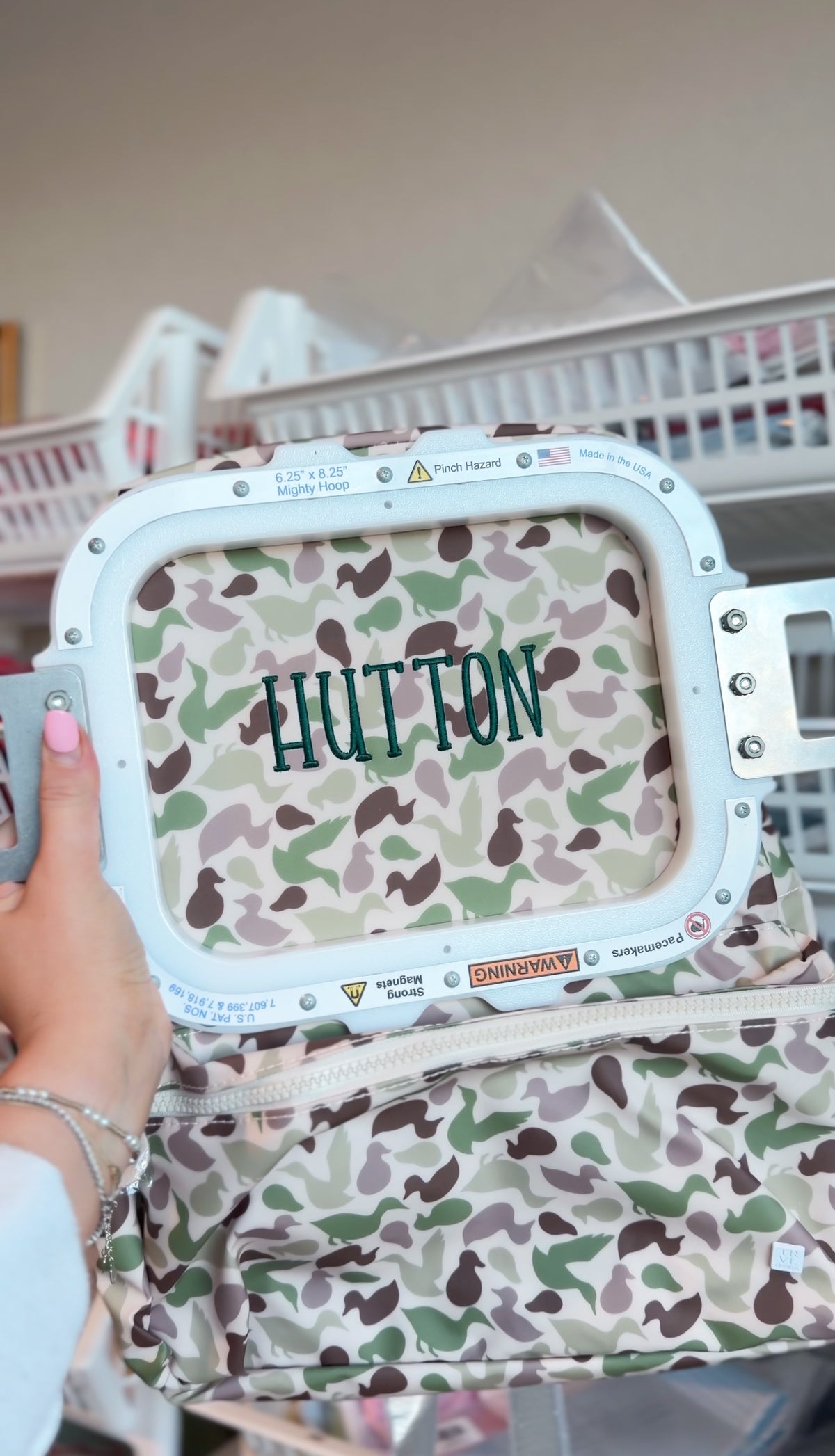 Camo duck print fabric being embroidered with the name 'Hutton' on an embroidery machine hoop