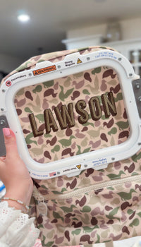 Camo duck print fabric being embroidered with the name 'Lawson' on an embroidery machine hoop