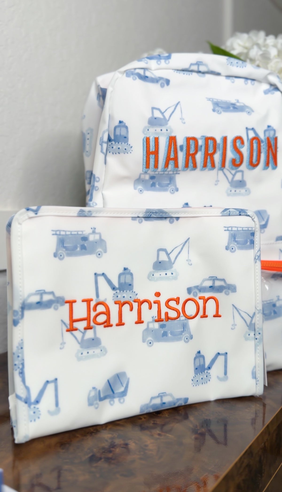 White TRVL pouch and backpacker with blue construction truck print, embroidered with the name 'Harrison' in orange