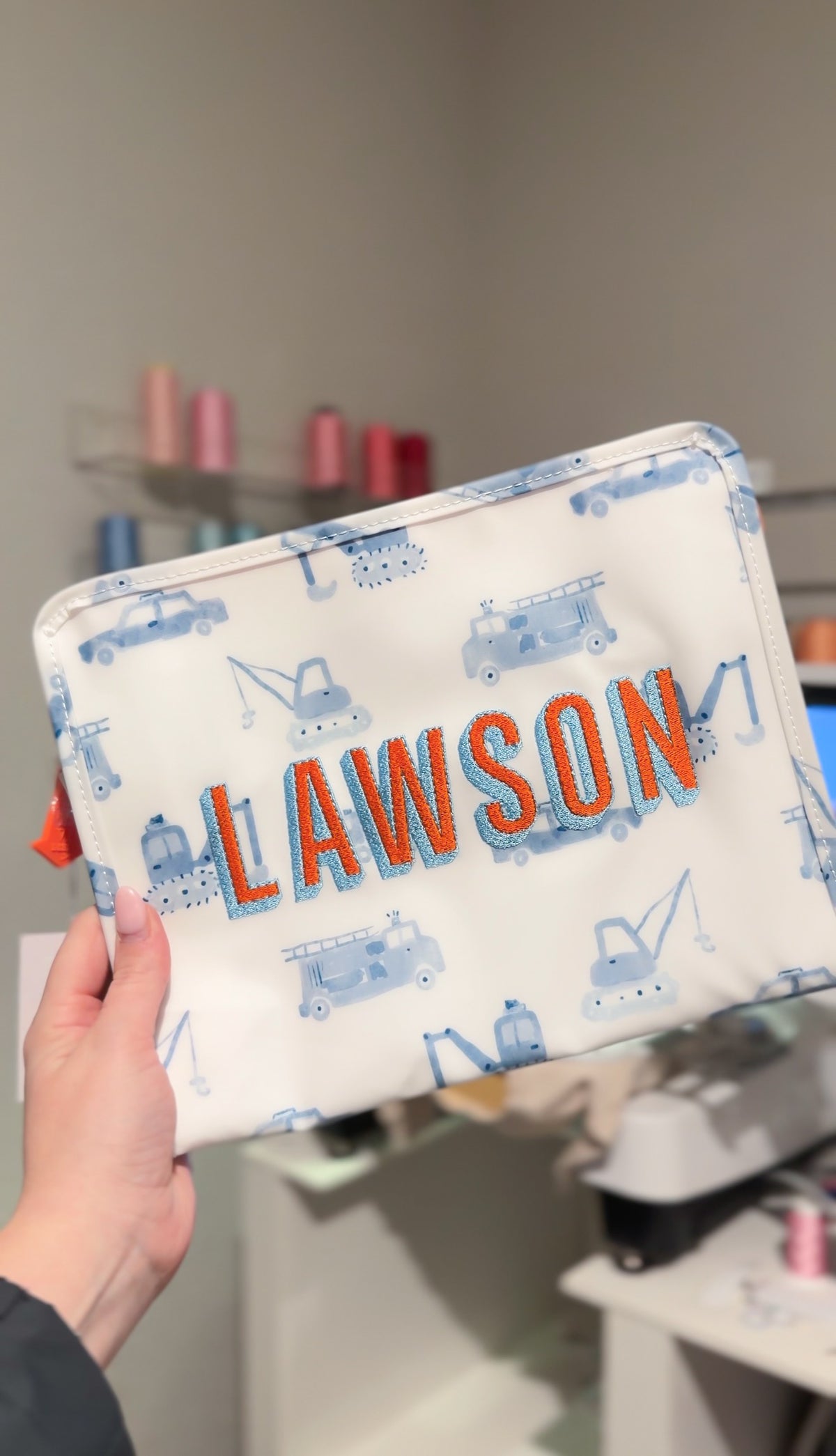 White TRVL pouch with blue construction truck print, embroidered with the name 'Lawson' in orange