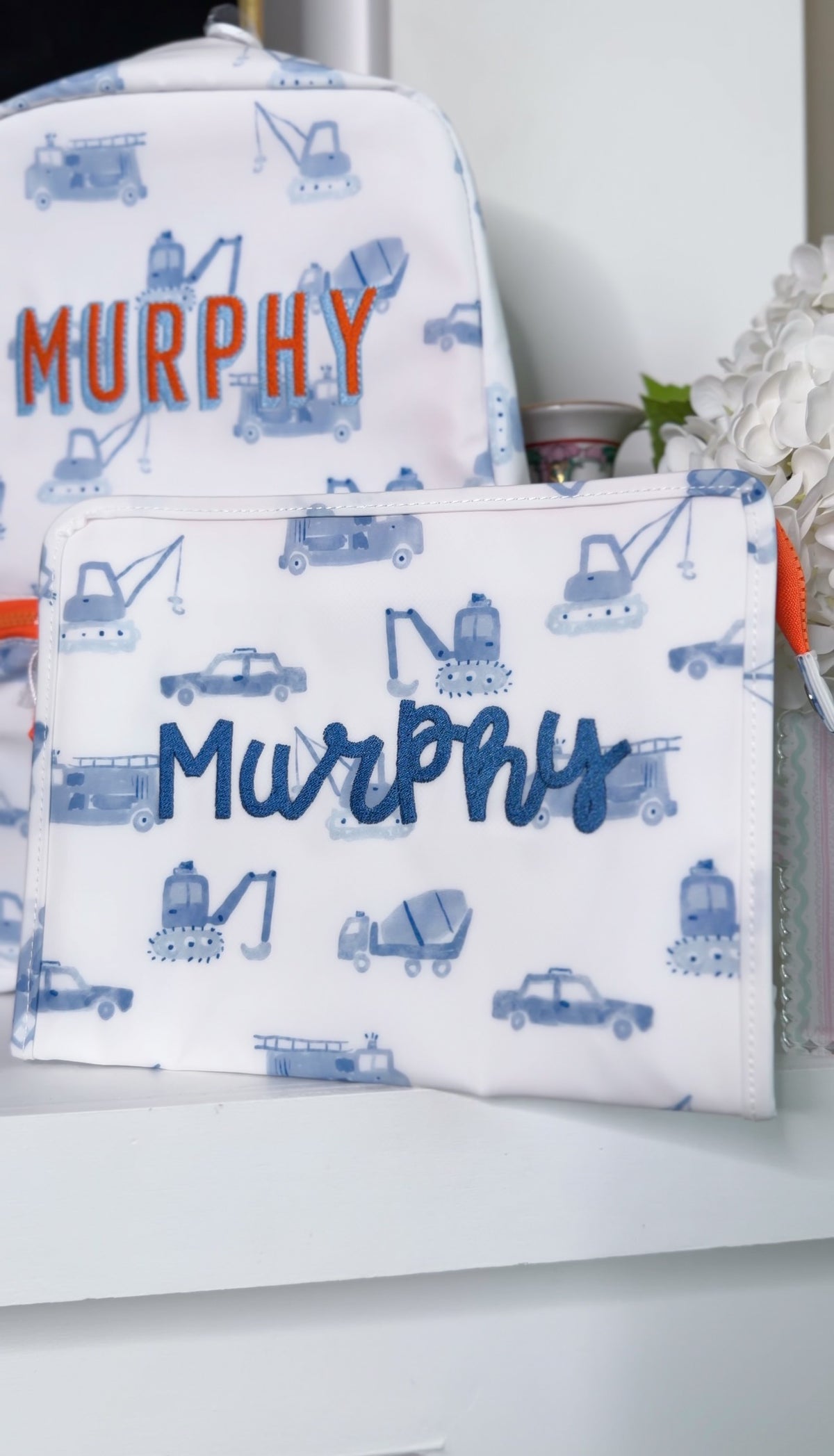 White TRVL backpack and pouch with blue construction truck print, embroidered with the name 'Murphy' in orange and blue