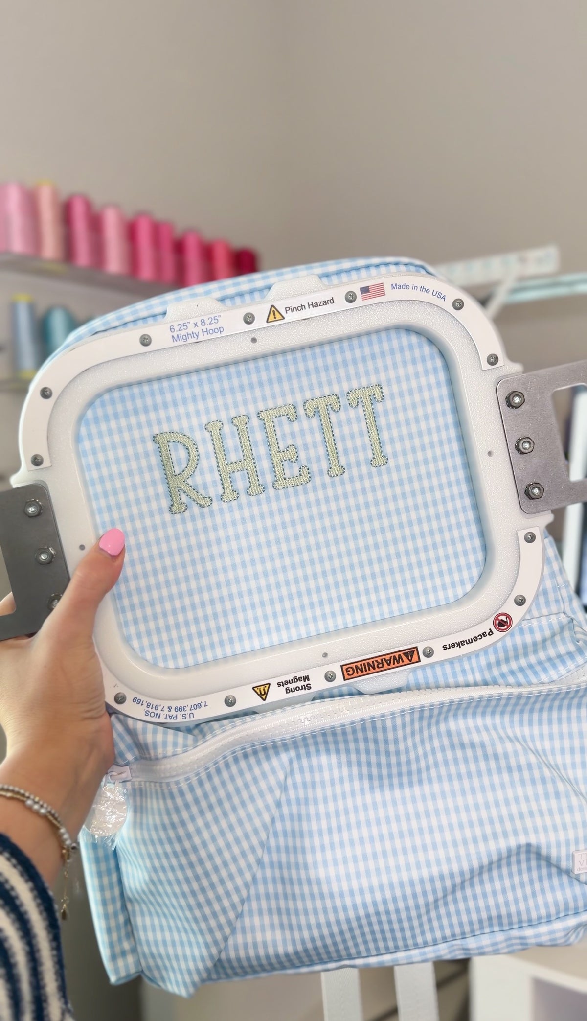 Light blue gingham fabric being embroidered with the name 'Rhett' on an embroidery machine hoop