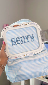 Light blue gingham fabric being embroidered with the name 'Henry' on an embroidery machine hoop