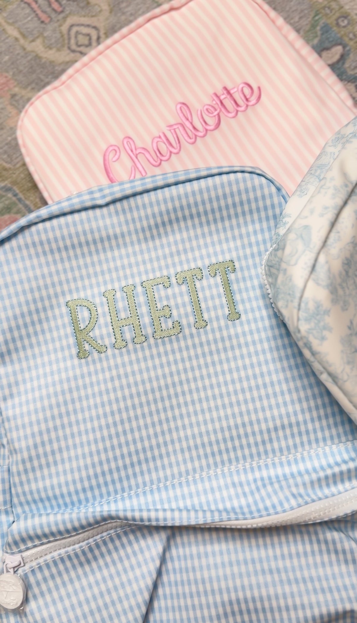 Light blue gingham TRVL Backpacker embroidered with the name 'Rhett', next to a pink striped bag embroidered 'Charlotte'