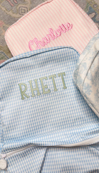 Light blue gingham TRVL Backpacker embroidered with the name 'Rhett', next to a pink striped bag embroidered 'Charlotte'