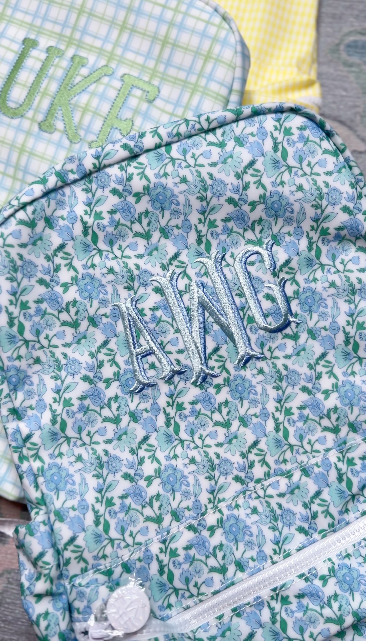Close-up of blue and green floral print TRVL Backpacker with light blue embroidered monogram, styled next to green plaid and yellow gingham bags