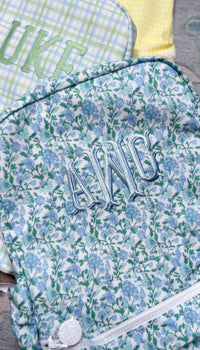 Close-up of blue and green floral print TRVL Backpacker with light blue embroidered monogram, styled next to green plaid and yellow gingham bags