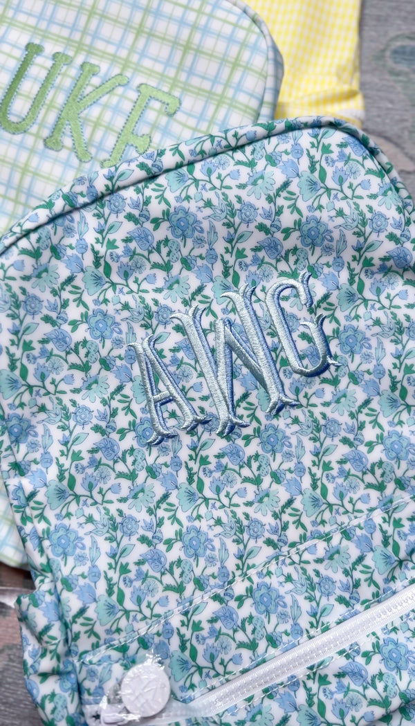 Close-up of blue and green floral print TRVL Backpacker with light blue embroidered monogram, styled next to green plaid and yellow gingham bags