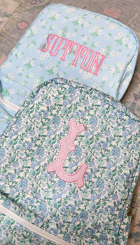 Blue and green floral TRVL Backpacker with pink embroidered 'L' monogram patch, next to a pale blue floral pouch embroidered 'Sutton'
