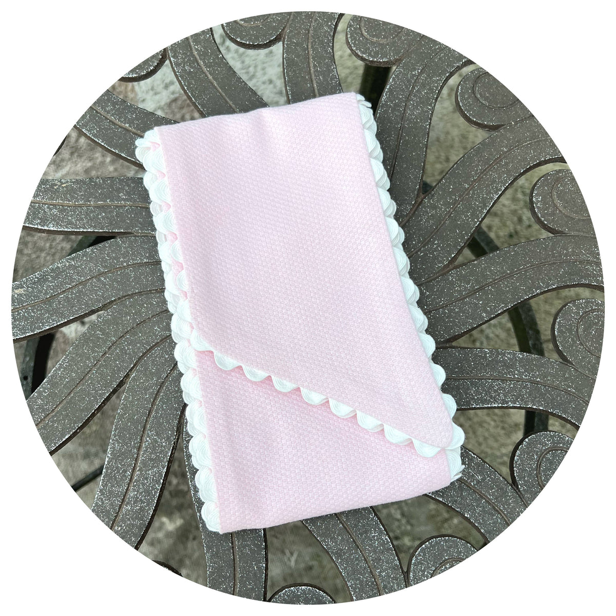 Folded pale pink cotton pique fabric with white scalloped ric rac trim, styled on an ornate metal outdoor table