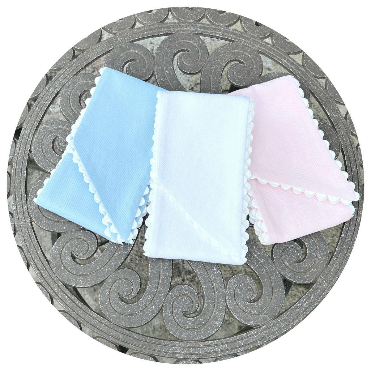 Three folded cotton pique sashes in light blue, white, and pink with white scalloped trim, arranged on a metal table