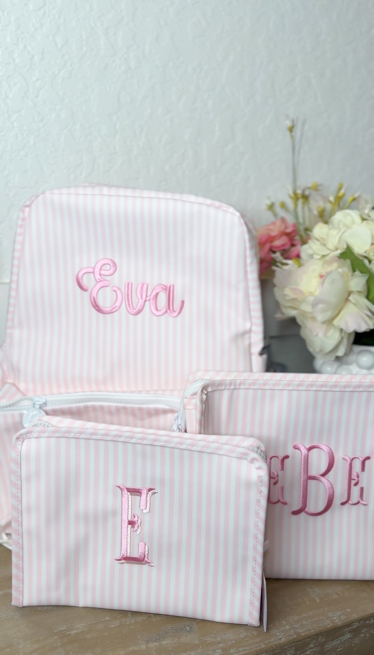 Pink and white striped backpack embroidered with the name 'Eva', next to matching pink striped pouches embroidered 'E' and 'B'