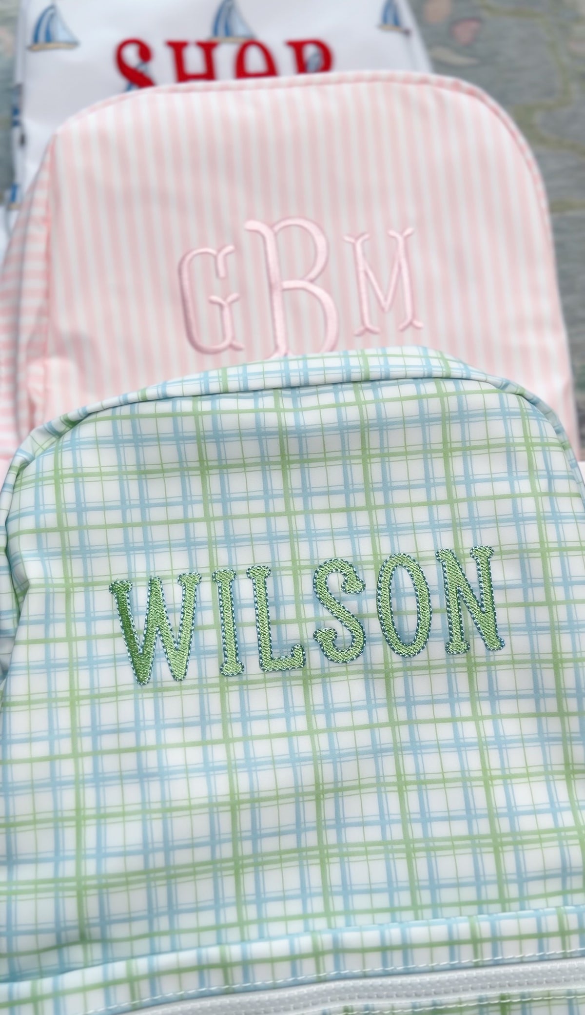 Pink and white striped backpack embroidered with a 'GBM' monogram, stacked with a sailboat print bag and a plaid backpack embroidered 'Wilson'