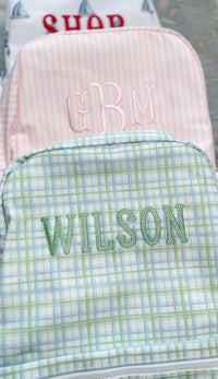 Pink and white striped backpack embroidered with a 'GBM' monogram, stacked with a sailboat print bag and a plaid backpack embroidered 'Wilson'