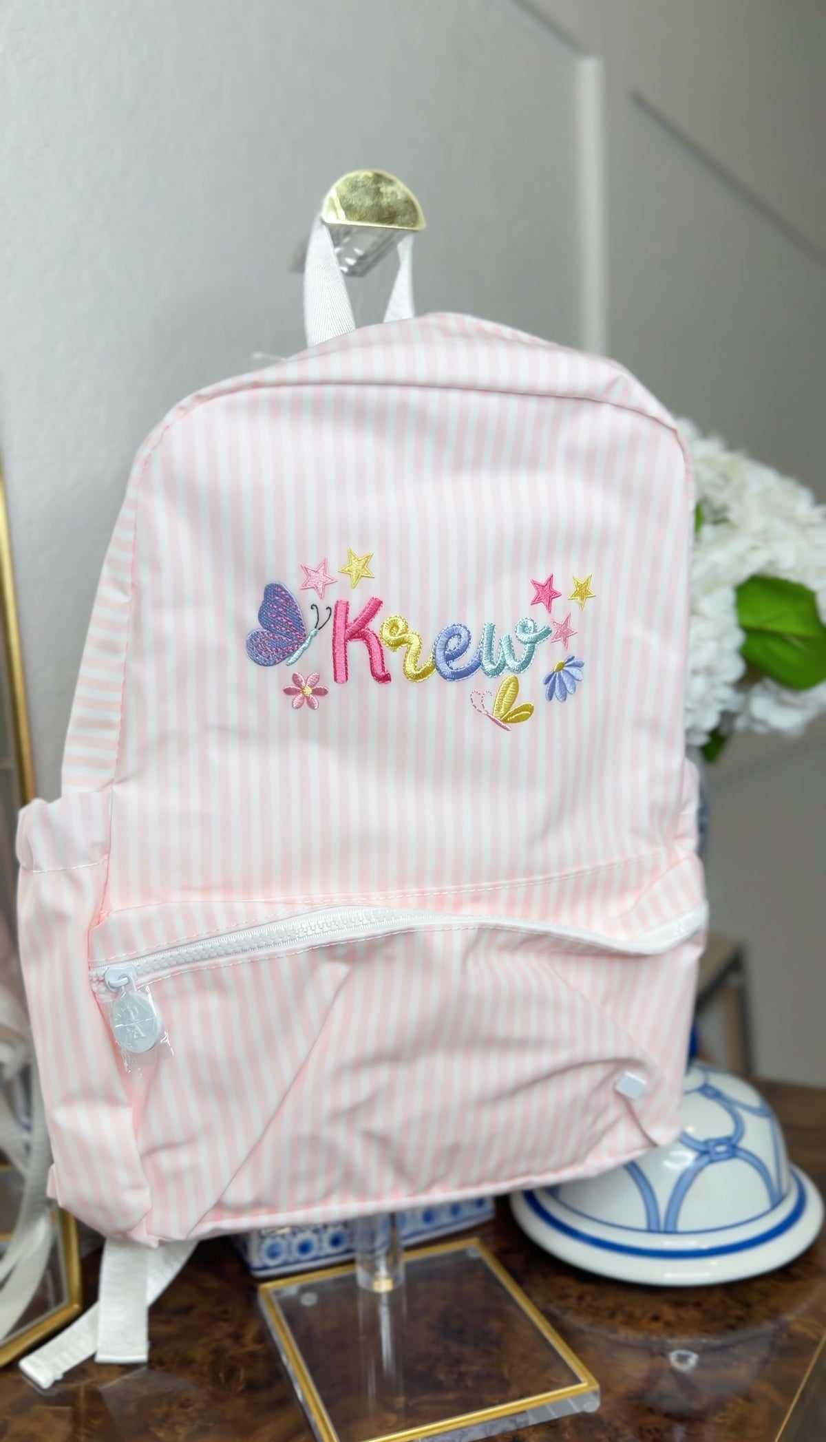 Pink and white striped backpack on display, embroidered with the name 'Krew' in colorful lettering with butterflies and stars