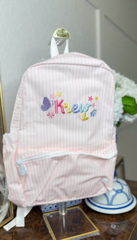 Pink and white striped backpack on display, embroidered with the name 'Krew' in colorful lettering with butterflies and stars