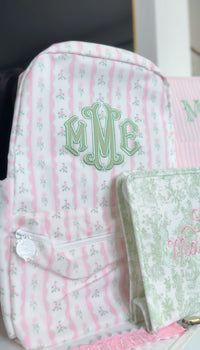Pink ribbon floral striped backpack embroidered with a green 'MME' monogram, next to a matching pouch