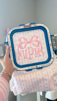 Pink and green floral embroidery hoop with a bow monogram 'VPM' design, held in front of a pink ribbon floral print bag