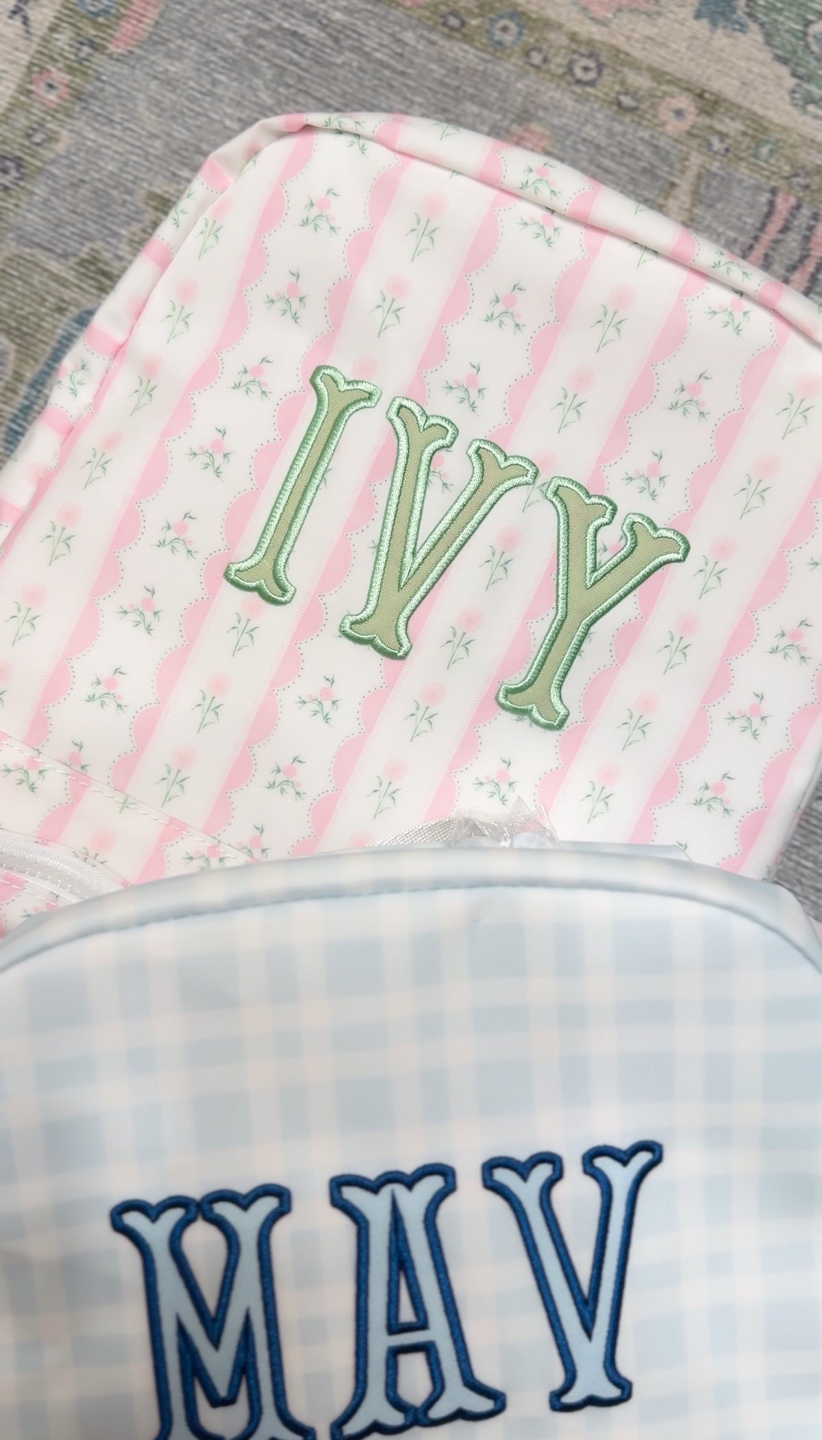 Pink ribbon floral striped bag embroidered with the name 'Ivy' in green, next to a blue gingham bag embroidered 'Mav'