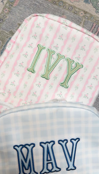 Pink ribbon floral striped bag embroidered with the name 'Ivy' in green, next to a blue gingham bag embroidered 'Mav'