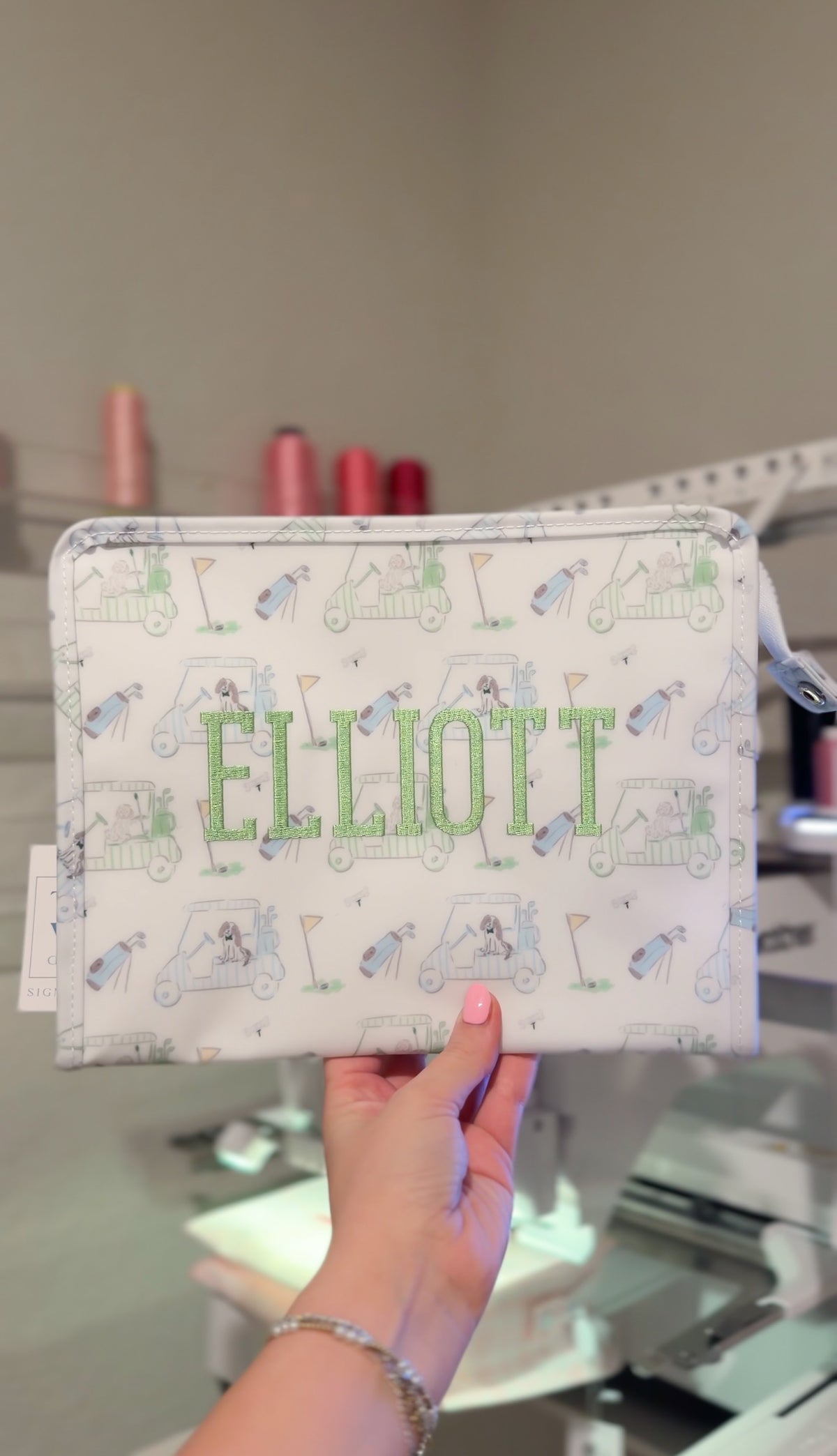 White TRVL pouch with green and blue golf print, embroidered with the name 'Elliott' in green