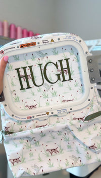 Green hunting dog print TRVL Backpacker being embroidered with the name 'Hugh' on an embroidery machine hoop