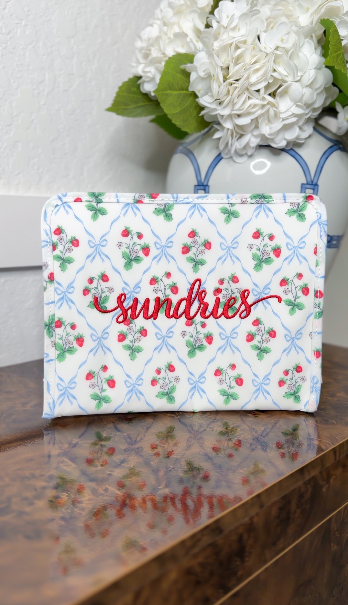 White TRVL pouch with red strawberry and blue bow trellis print, embroidered 'sundries' in red script