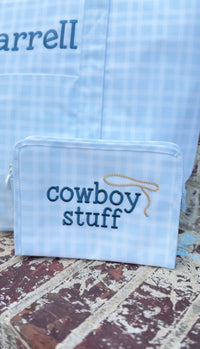 Light blue plaid TRVL pouch embroidered 'cowboy stuff' in navy, next to a bag embroidered 'arrell'