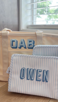 Blue and white striped TRVL Roadie pouch embroidered with 'OWEN' in bold block letters, shown with a matching tote bag