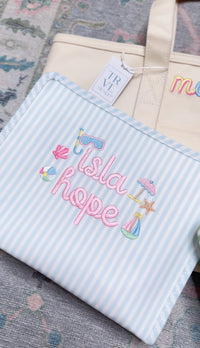Blue Pimlico stripe TRVL Roadie pouch embroidered with 'Isla Hope' and beach icons, resting against a cream tote bag