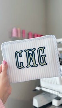 Blue Pimlico stripe TRVL Roadie pouch embroidered with 'CWG' monogram, held next to an embroidery machine