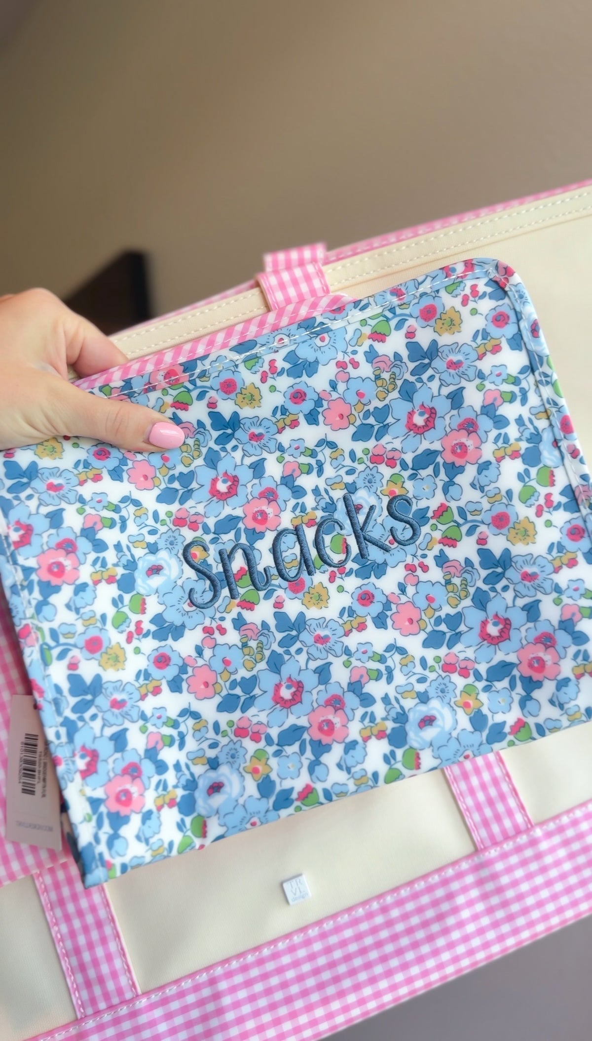 Blue floral TRVL pouch embroidered 'Snacks' in navy script, held next to a pink gingham bag