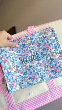 Blue floral TRVL pouch embroidered 'Snacks' in navy script, held next to a pink gingham bag